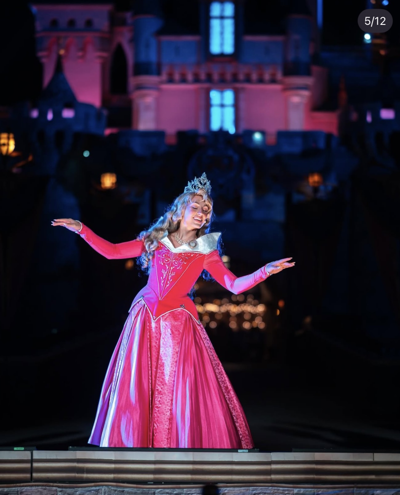Princess in pink gown with crown, arms outstretched, in front of castle backdrop at night.