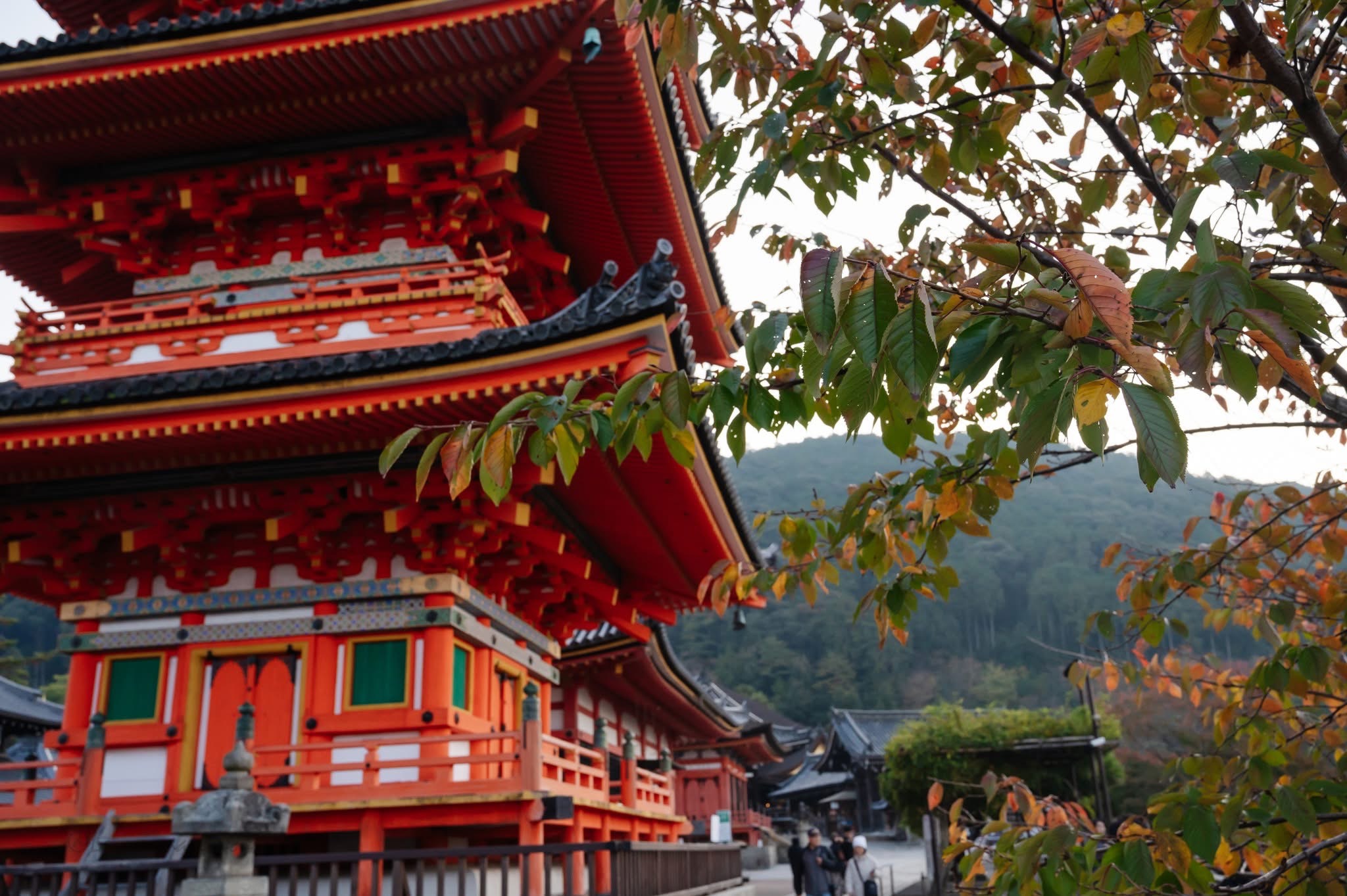 Traditional multi-tiered red and orange temple with intricate details, surrounded by green and orange leaves, mountains in background.