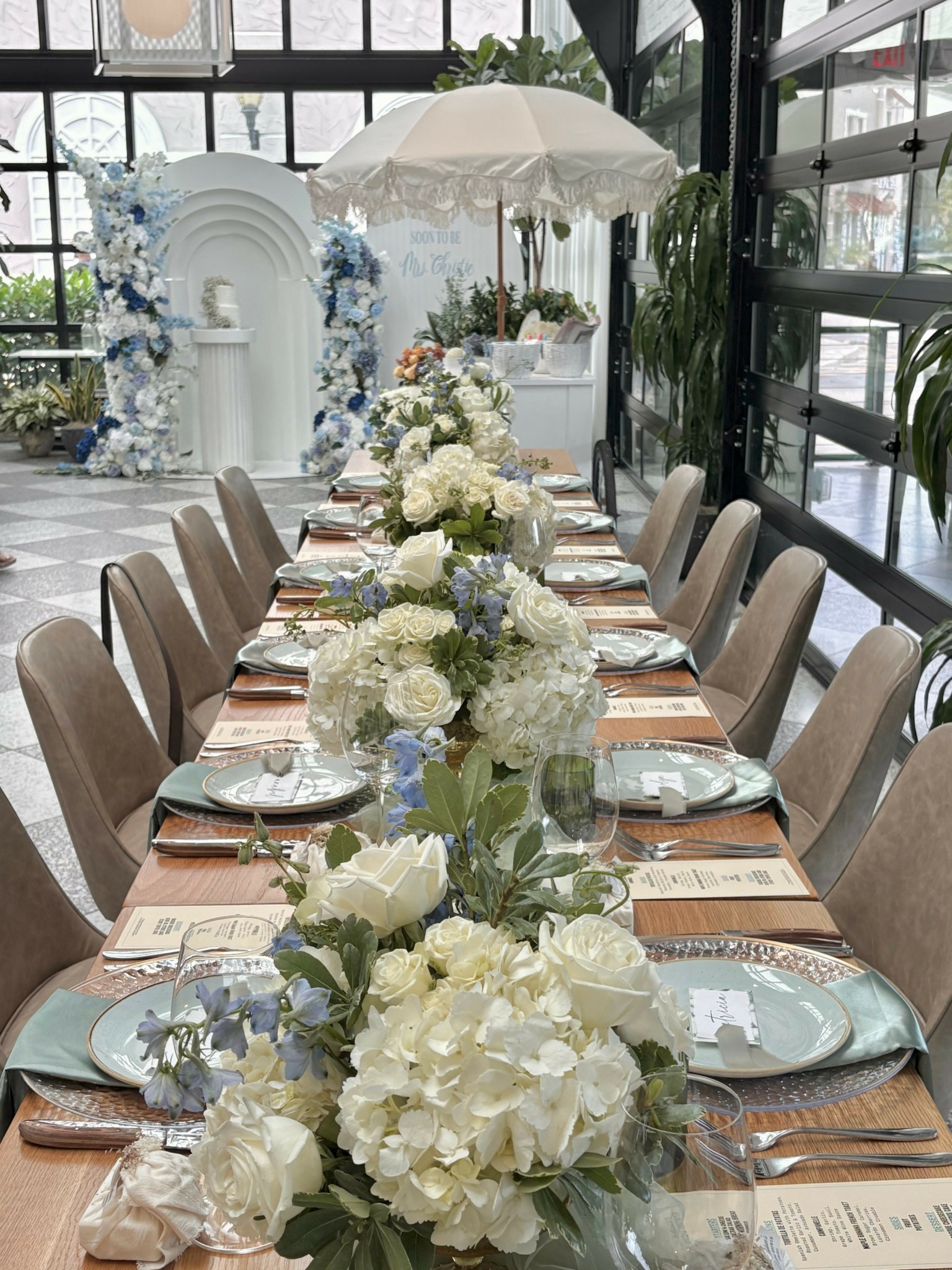 Long dining table with floral centerpieces, set with plates, glasses, and napkins, in a bright indoor space with large windows.