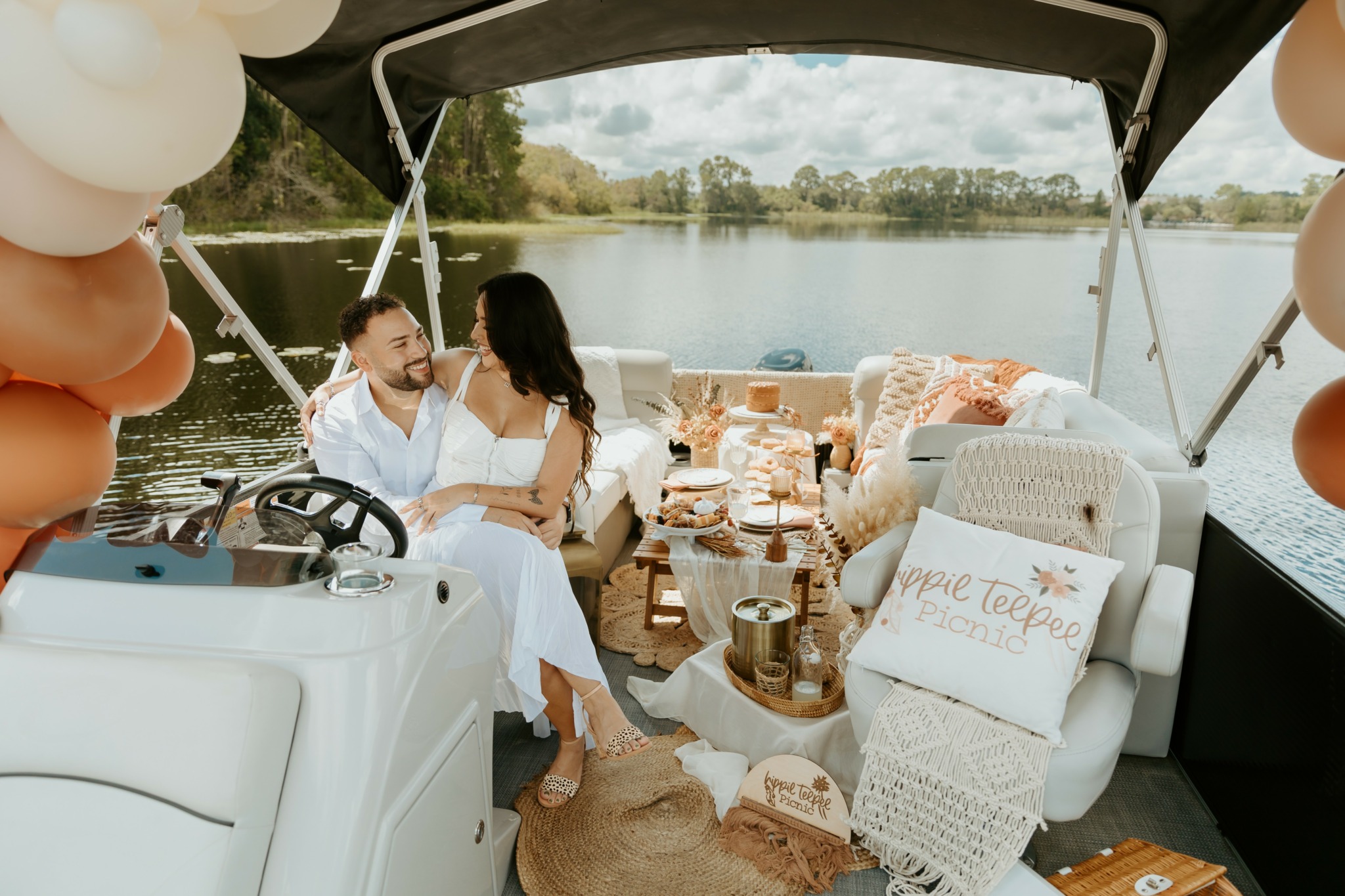 Two people sit on a boat near a lake, surrounded by balloons and decorations, with a table and seating area.