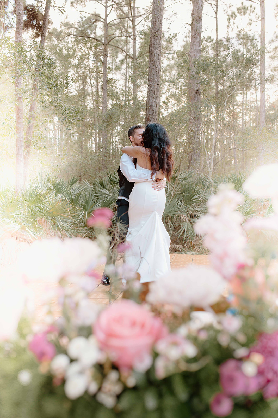 A couple embraces in a forest with tall trees, surrounded by pink and white flowers, sunlight filtering through the trees.