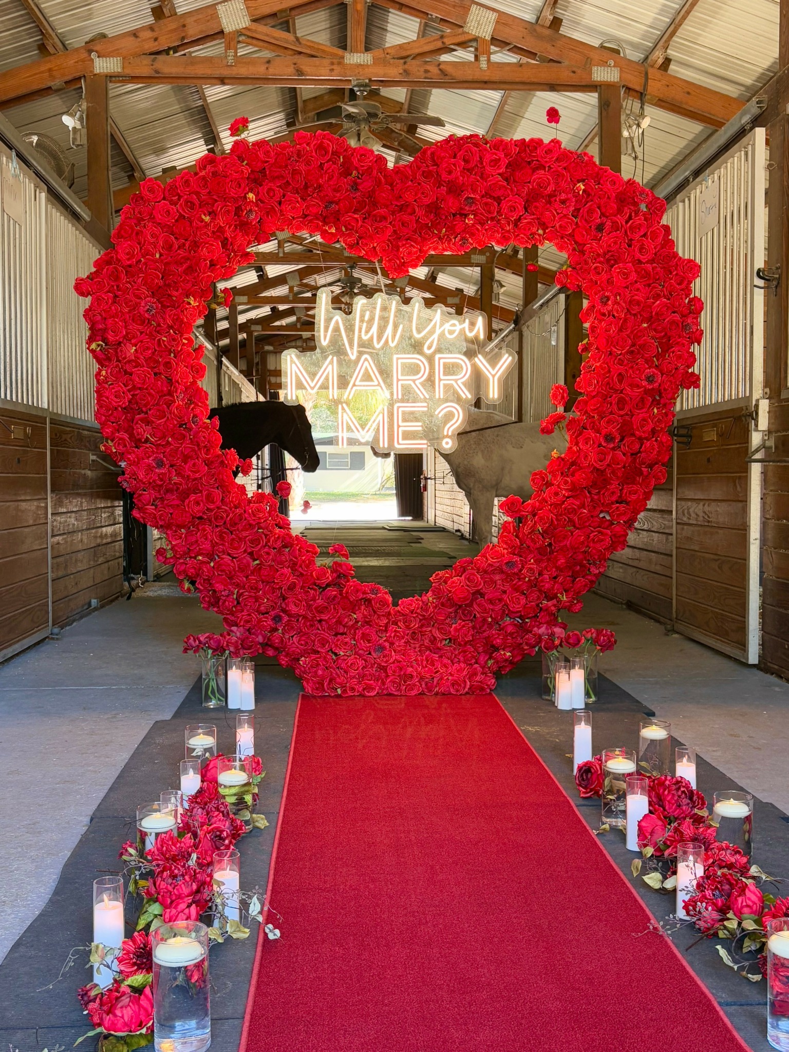 Heart-shaped floral arch with red flowers, red carpet, and candles along the aisle, inside a wooden structure.