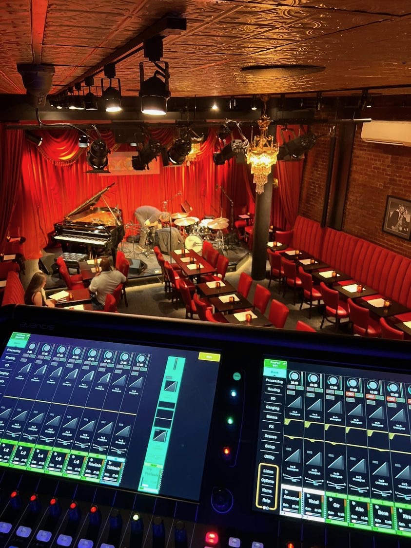 Jazz band performing on stage with red curtains, audience seated at tables, dim lighting, and a chandelier, viewed from sound mixing console.