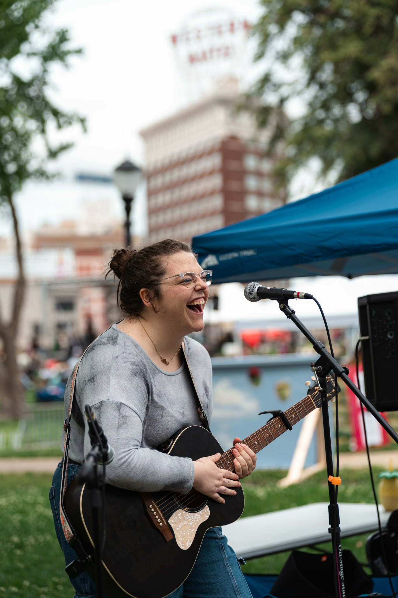 Woman singing and playing guitar outdoors near a blue tent, with a blurred cityscape background.