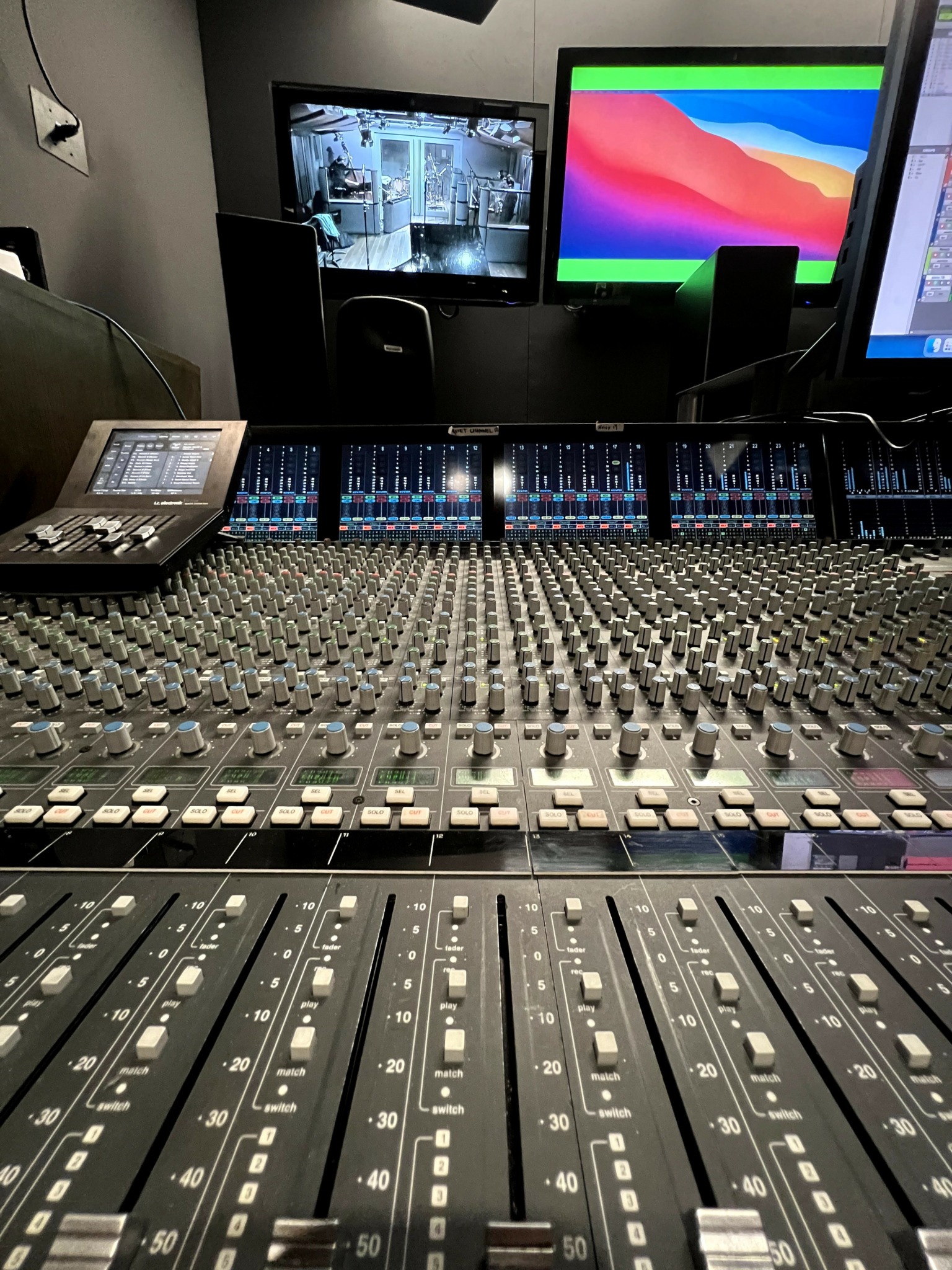 Recording studio control room with mixing console, monitors, and equipment, viewed from the mixing desk, with screens displaying different visuals.
