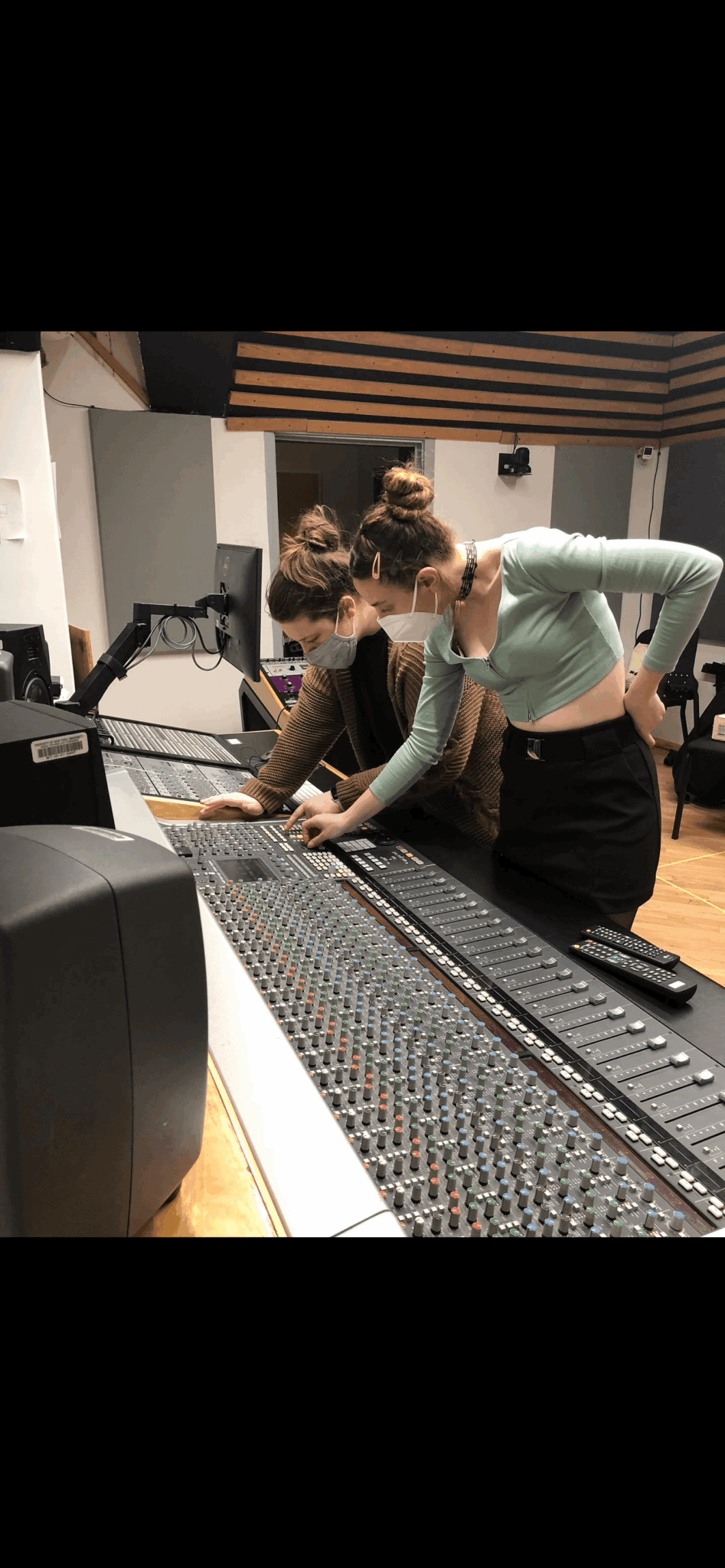 Two women work together at a large audio mixing console in a recording studio, with equipment and soundproofing visible.