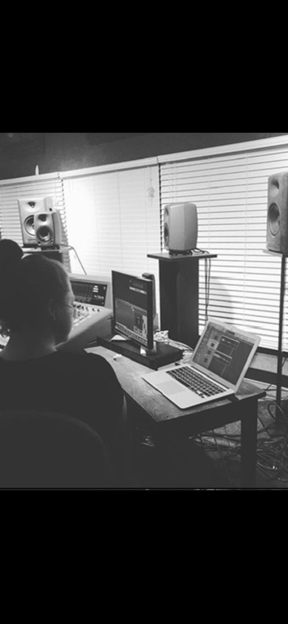 Person working at a computer in a music studio with speakers and equipment, window blinds in the background.