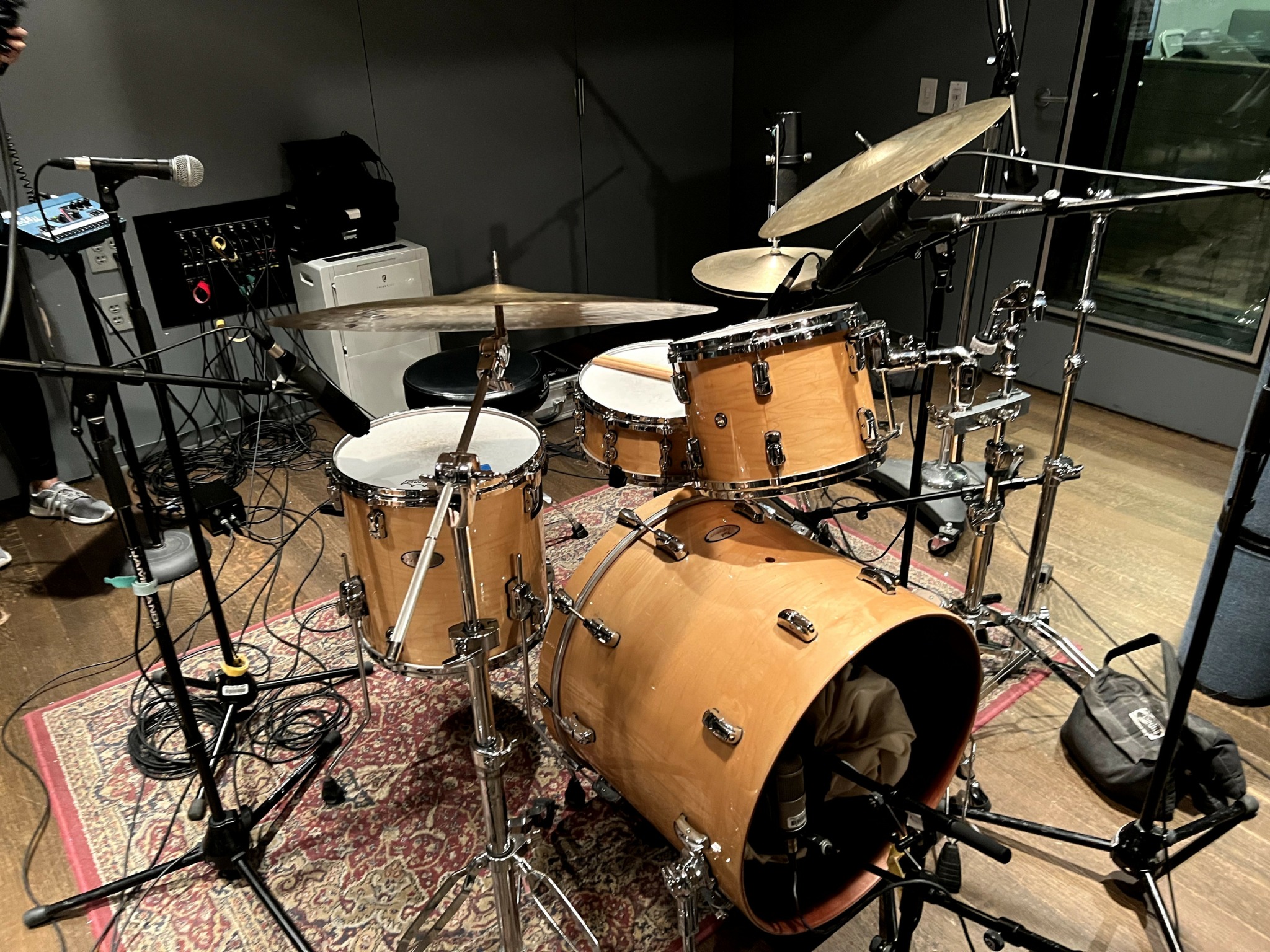 A drum set with multiple drums, cymbals, and microphones in a recording studio, with a window and equipment in the background.