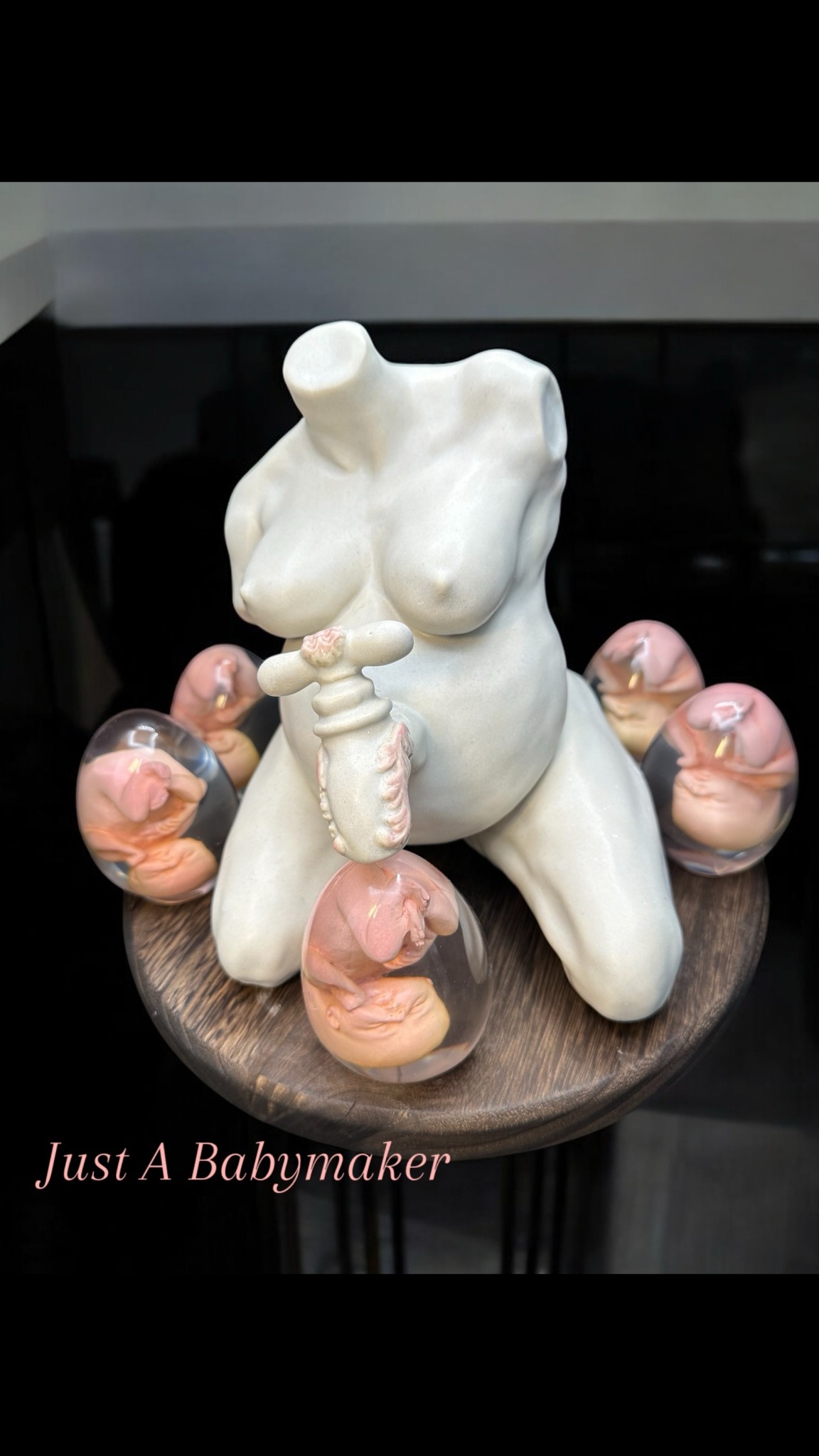 Sculpture of a female torso with multiple baby figurines surrounding it on a round wooden base, with text 