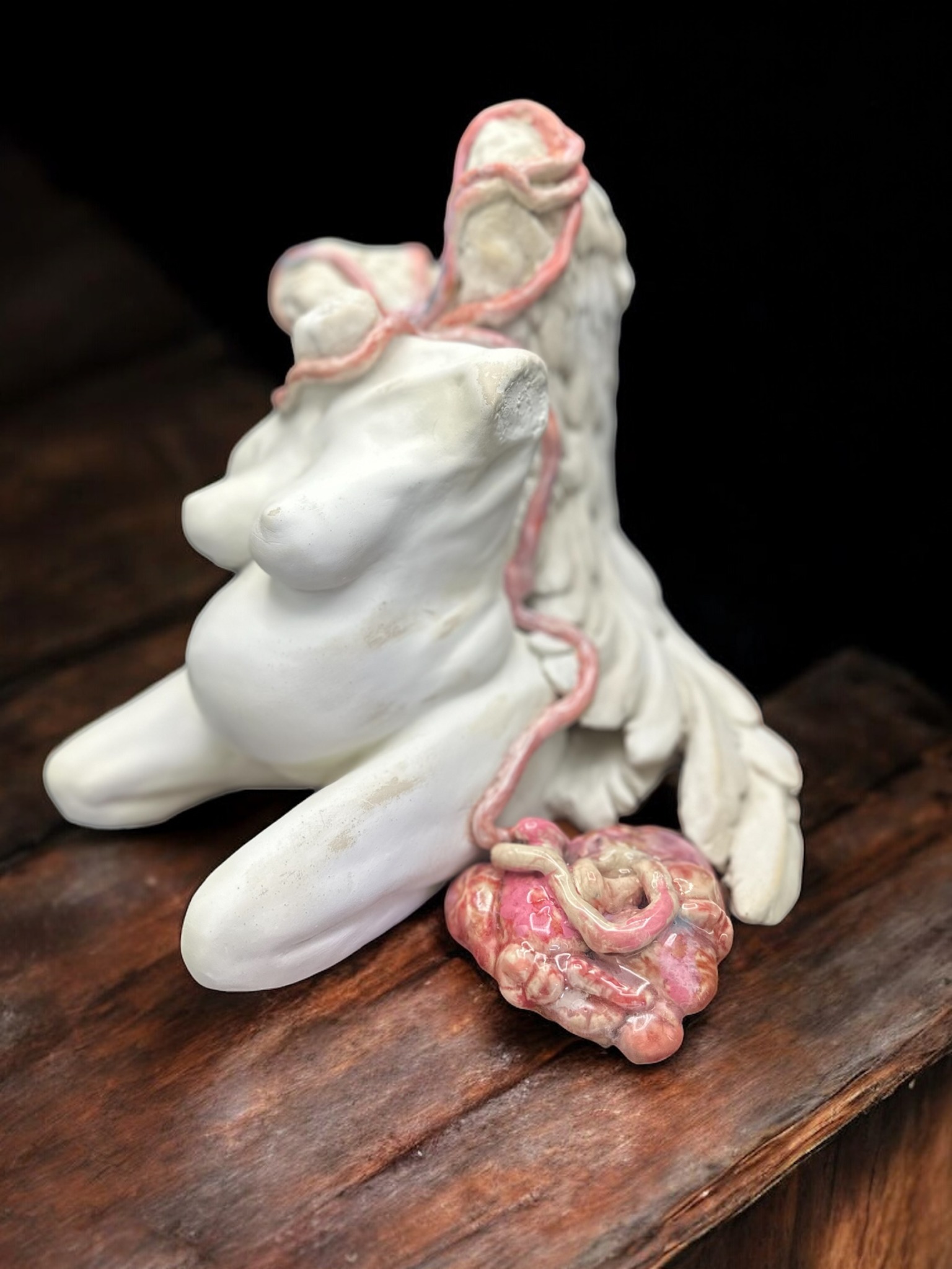 Ceramic sculpture of a seated figure with a pink, textured object attached, on a wooden surface.