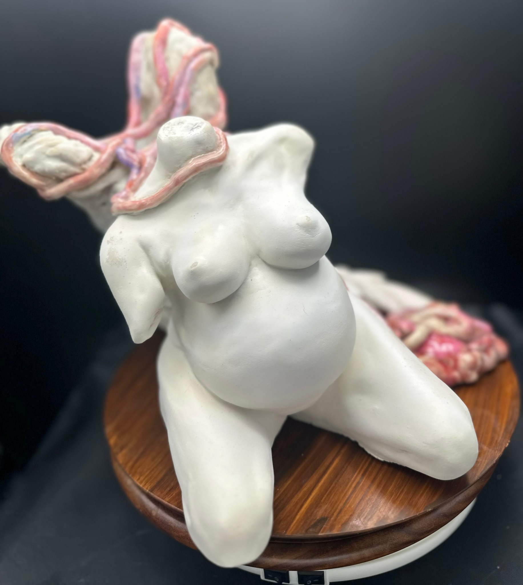 A sculpture of a female torso with a rabbit head, placed on a wooden base, against a dark background.