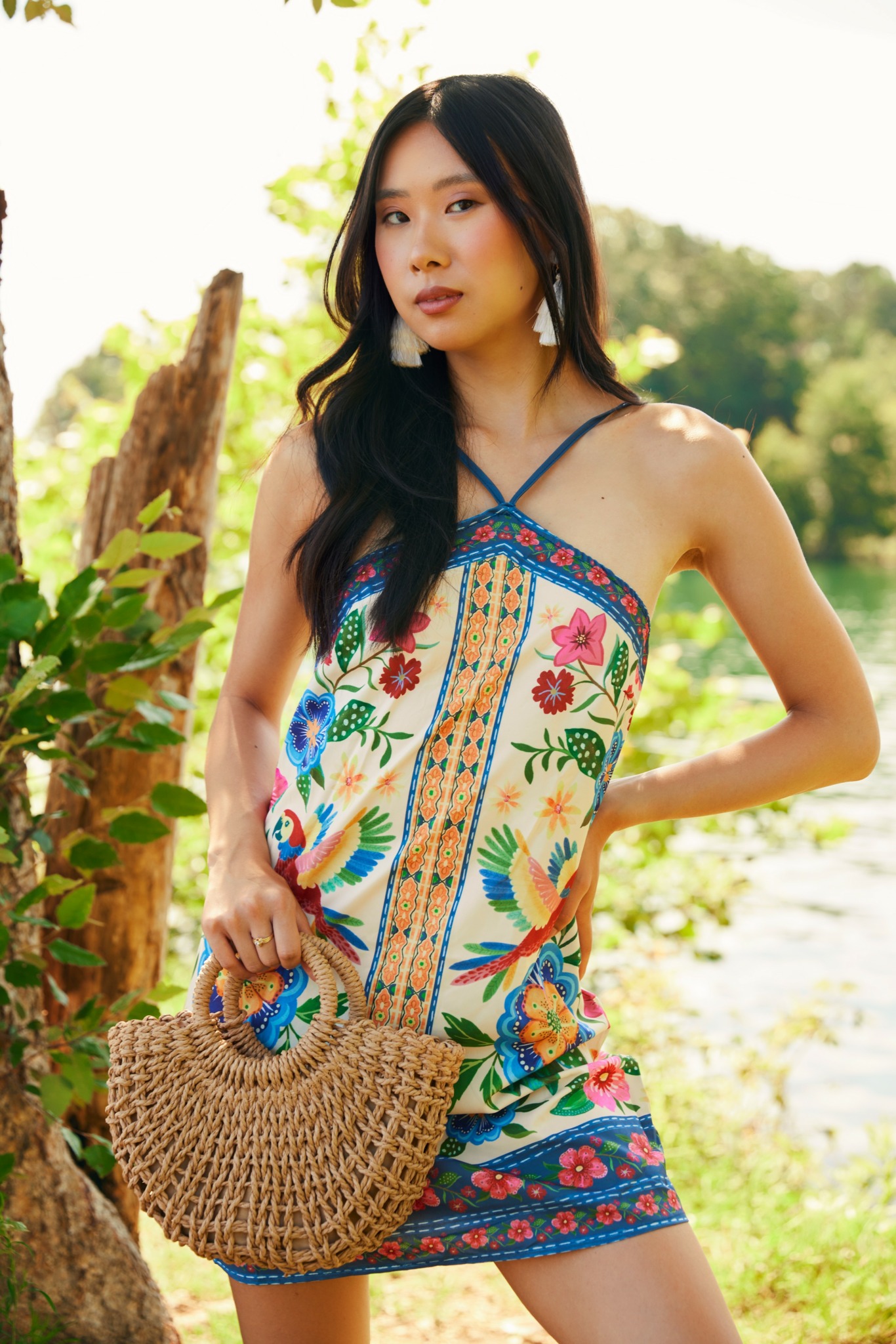 Young woman in colorful dress holding a woven bag outdoors, with trees and water in background.