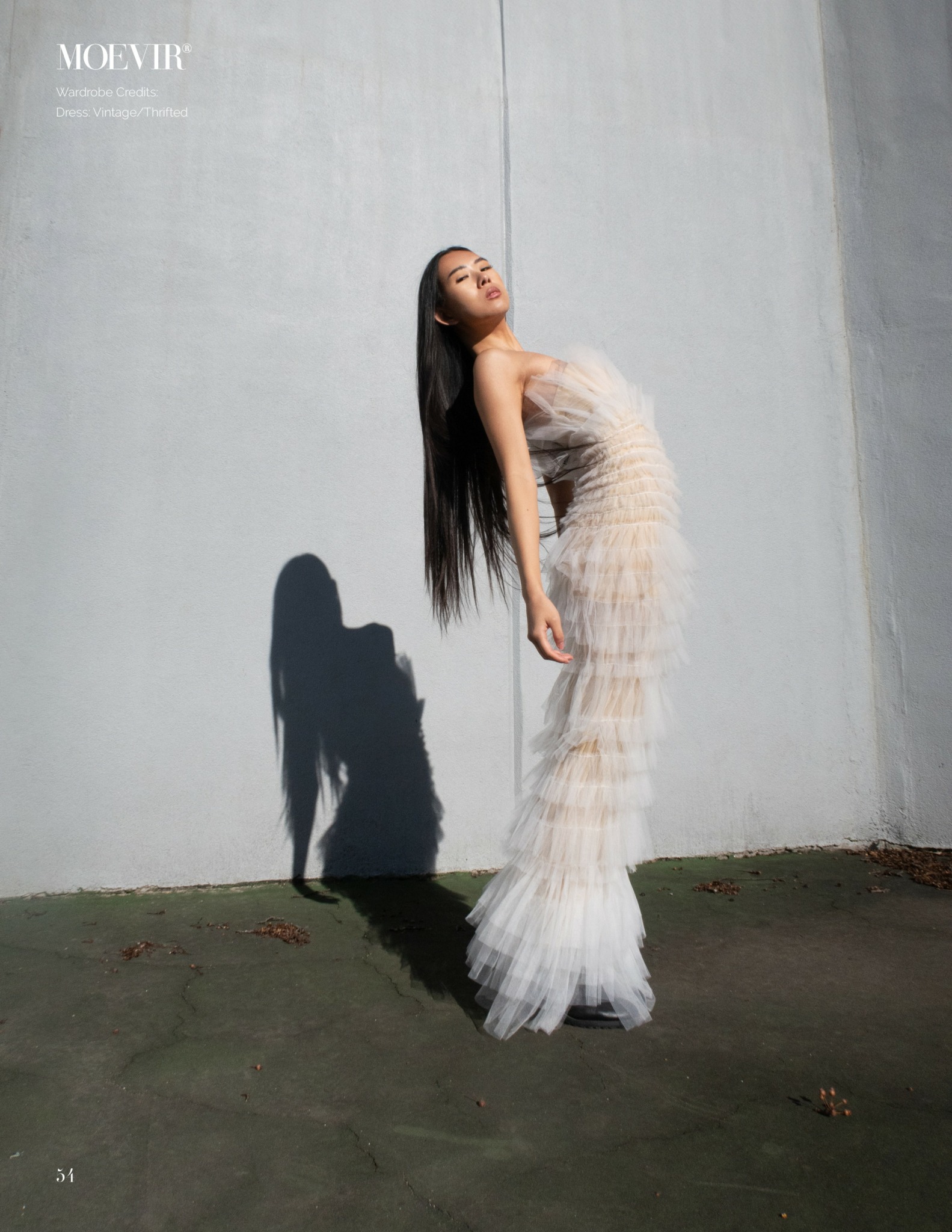 Woman in a strapless, layered dress standing against a plain wall, casting a shadow on the ground.