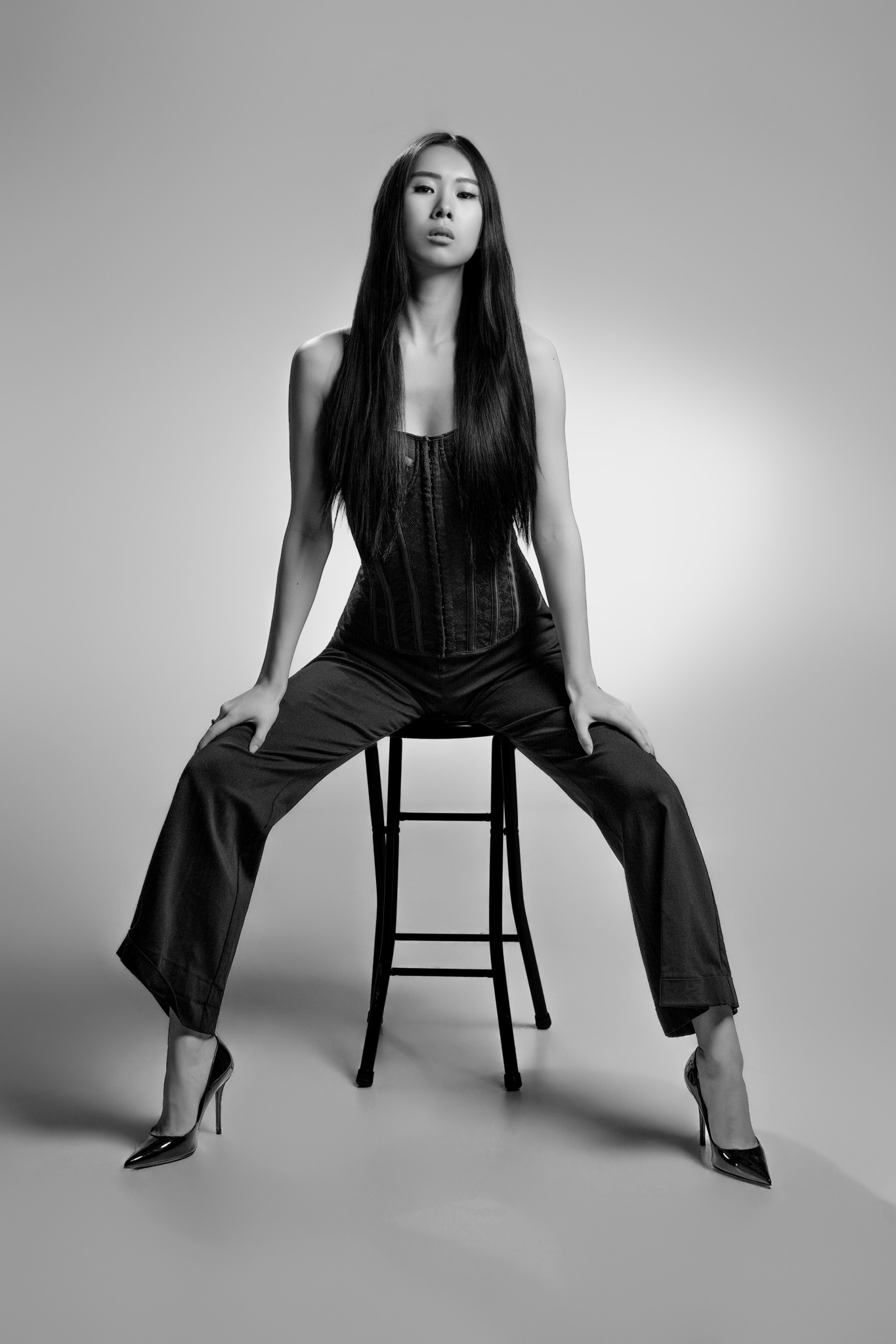 Woman with long hair sitting on a stool, legs apart, in a black outfit, in a studio setting.