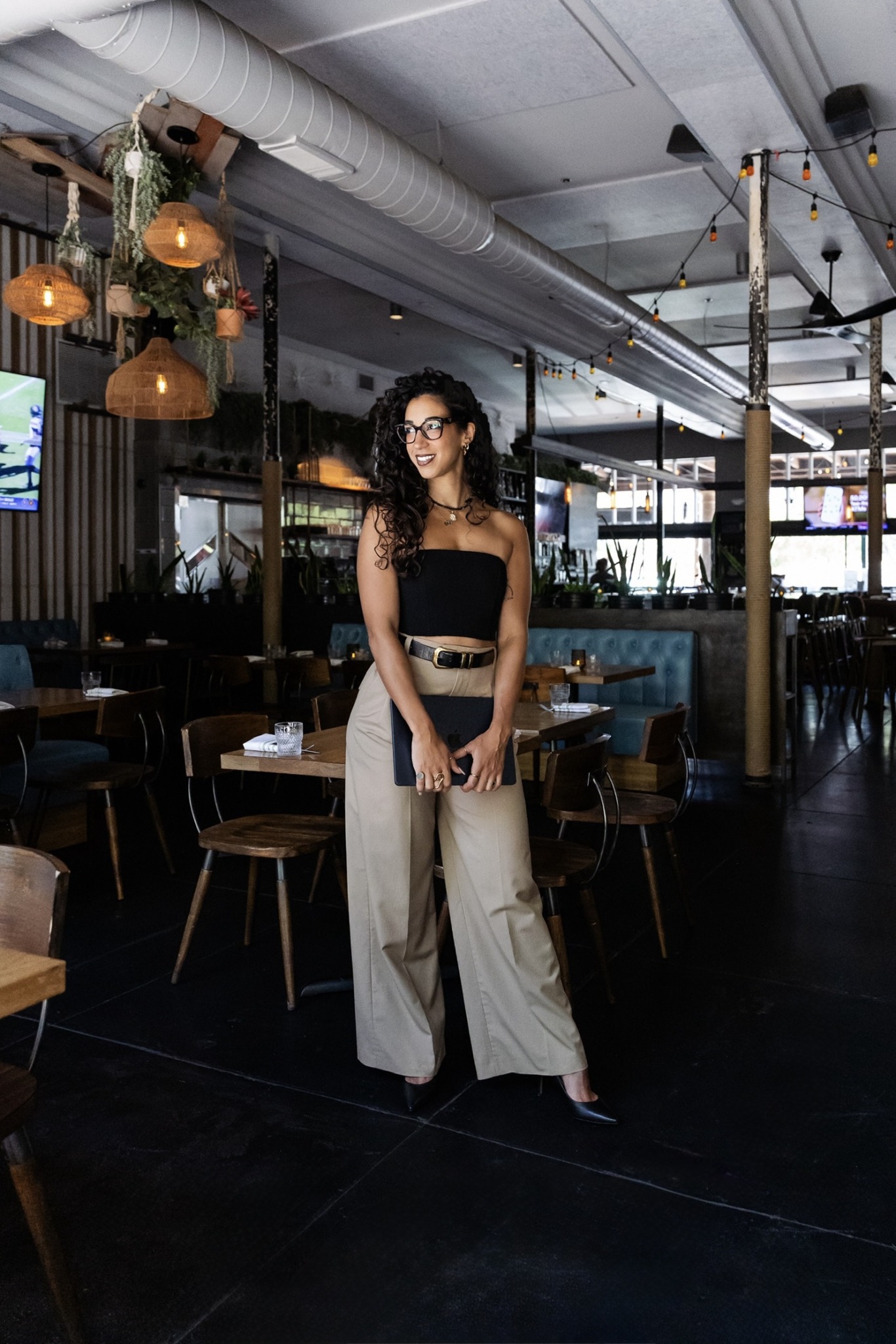 Woman with curly hair and glasses standing in a modern restaurant with hanging lights and large windows.