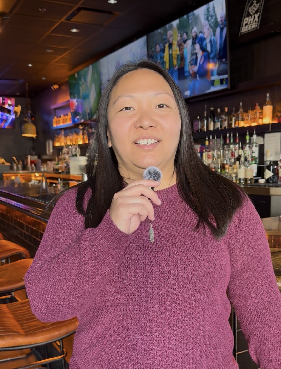 Woman in pink sweater holding a lollipop inside a bar with bottles and TV screens behind her.