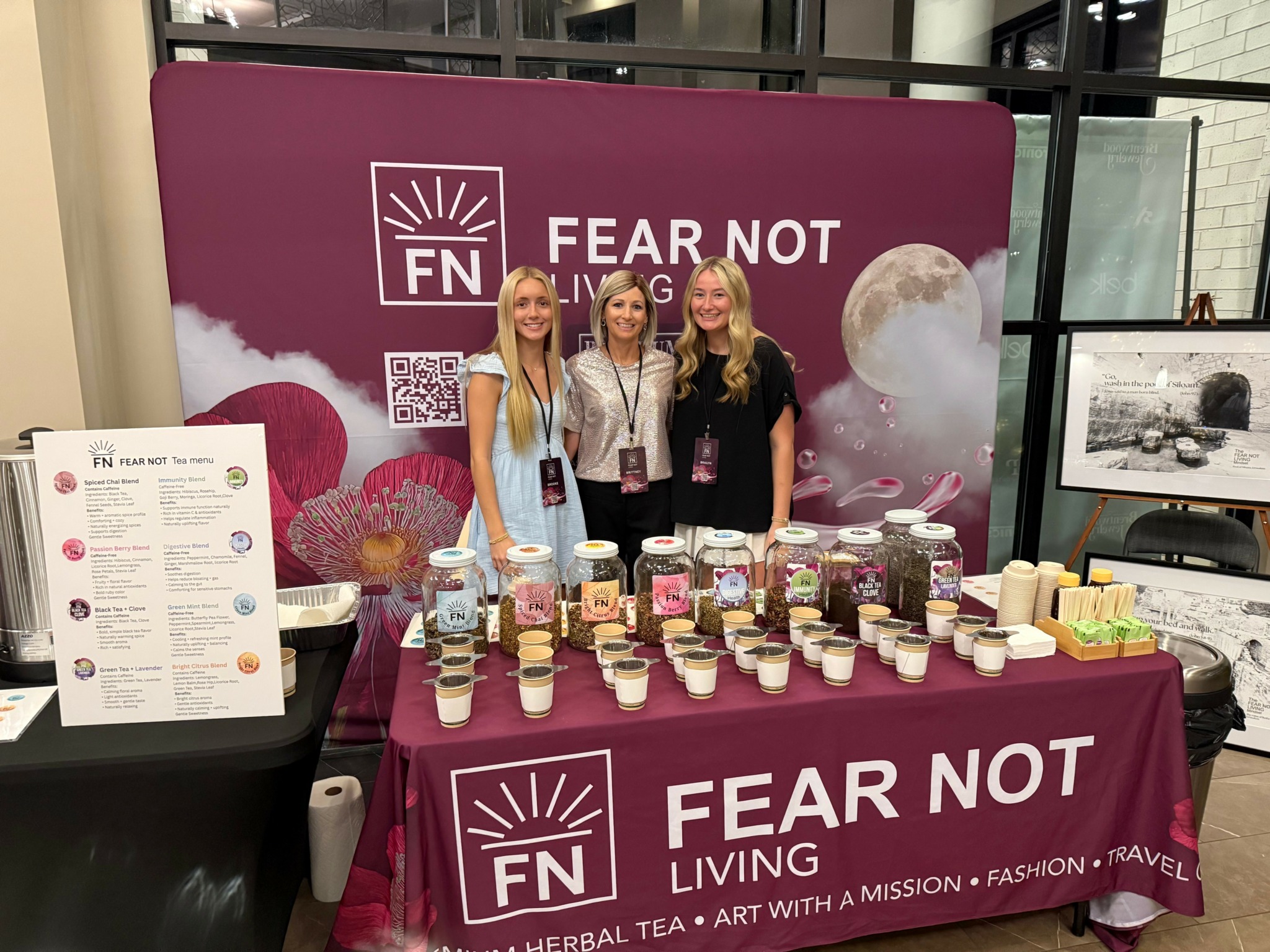 Three women stand behind a table with jars and soap bars at a promotional booth, with a purple backdrop reading 'FEAR NOT LIVING' and 'FN'.