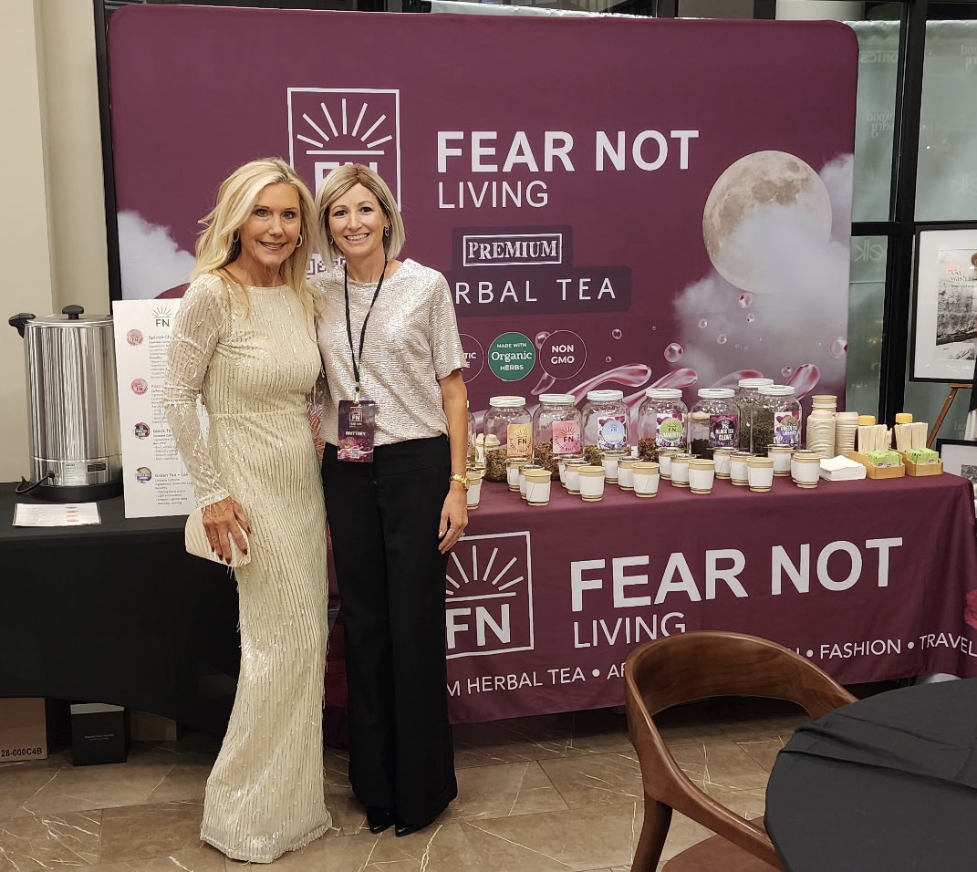 Two women standing in front of a purple display booth with tea products, smiling, one in a cream dress, the other in black pants and a patterned top.