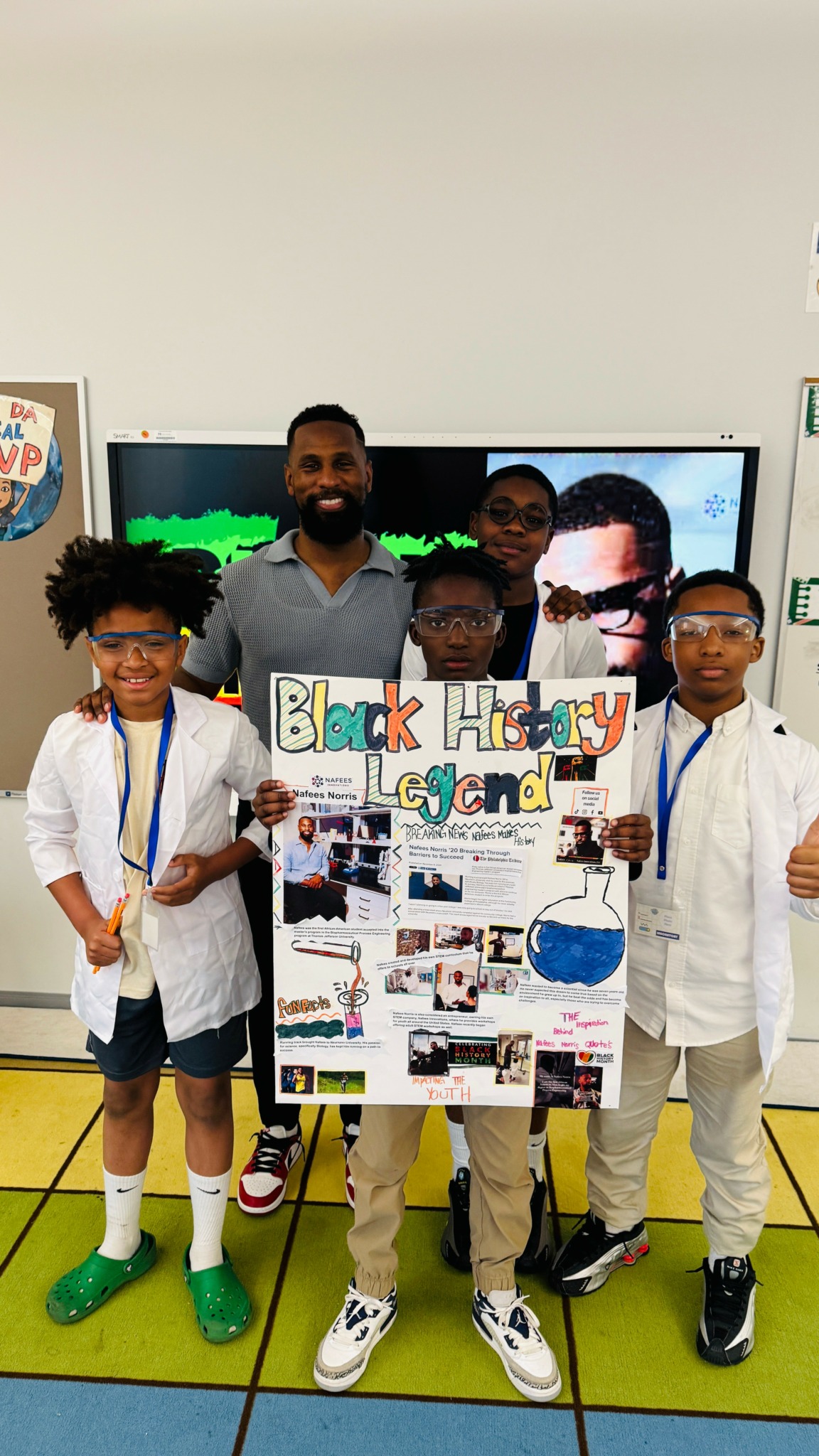 Group of five children and one adult holding a poster in a classroom, with a screen displaying a person's face behind them.