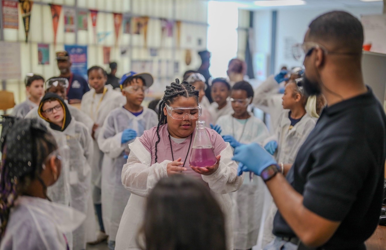 Group of children in lab coats gathered around a person holding a bowl, in a science classroom or lab.
