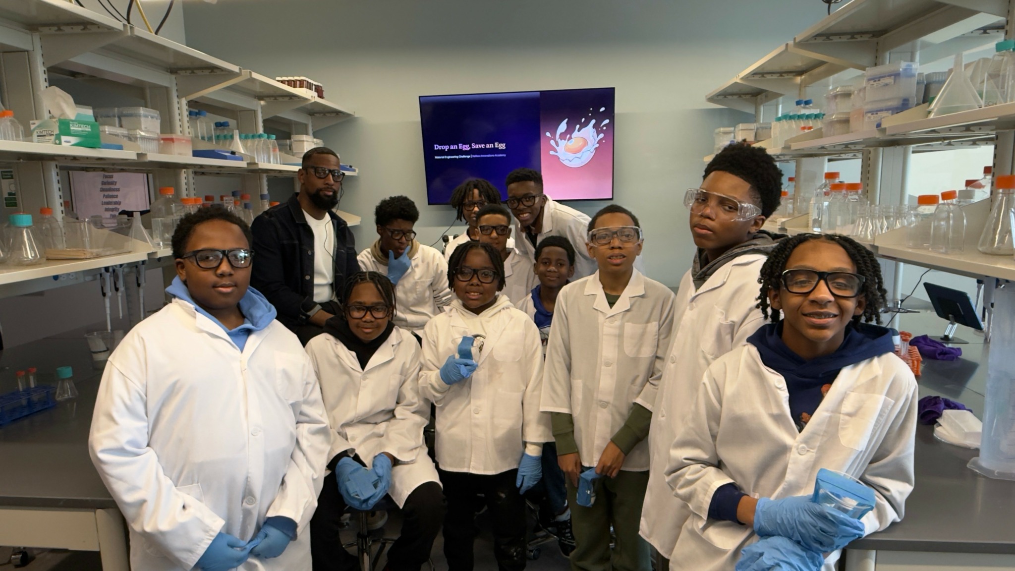 Group of people in a laboratory wearing white lab coats and gloves, with shelves and a screen in the background.