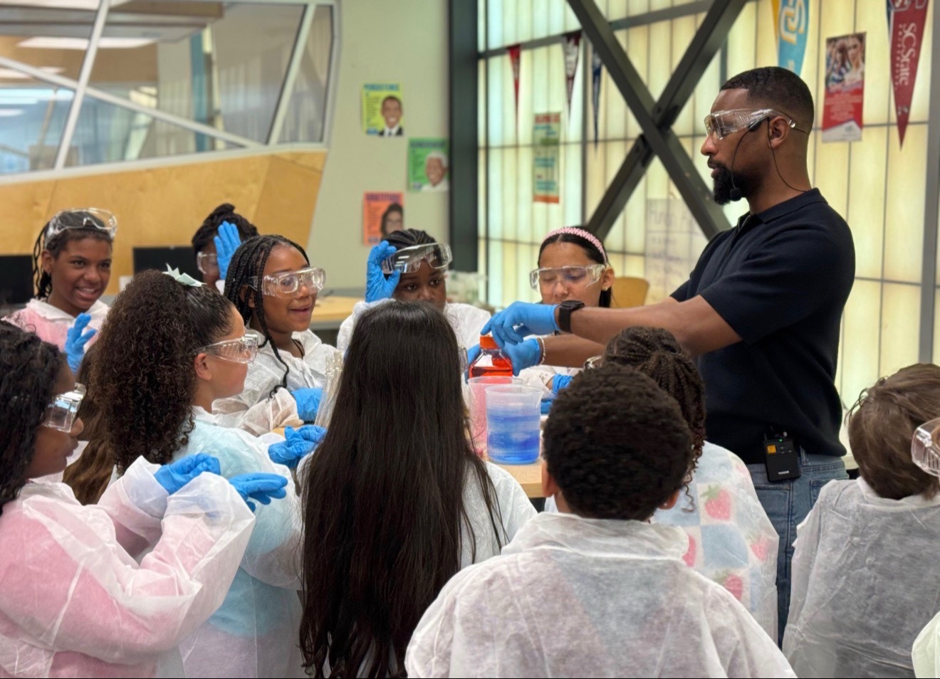 Man instructing children in a science activity, children wearing lab coats and gloves, colorful classroom background.