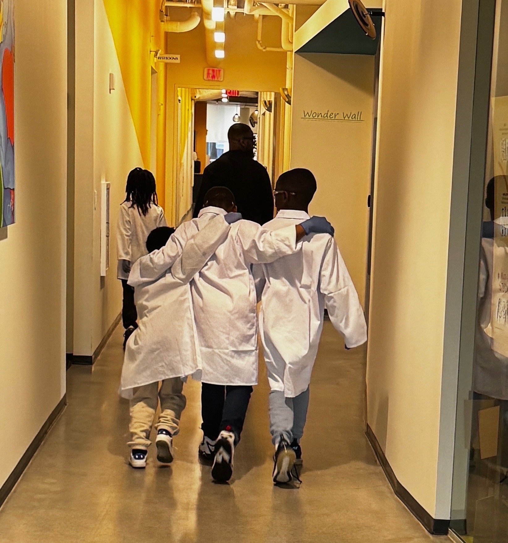 Three children in white coats walk down a brightly lit yellow hallway with a person ahead, and a girl in a white jacket behind.