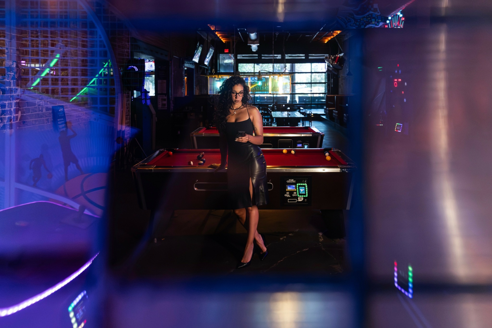 Woman in black dress standing near pool table in dimly lit bar with neon lights, windows in background.