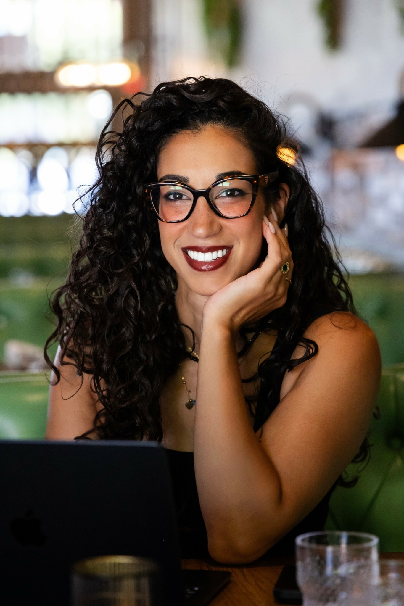 Woman with curly hair and glasses smiling, resting her chin on her hand, in a cafe setting.