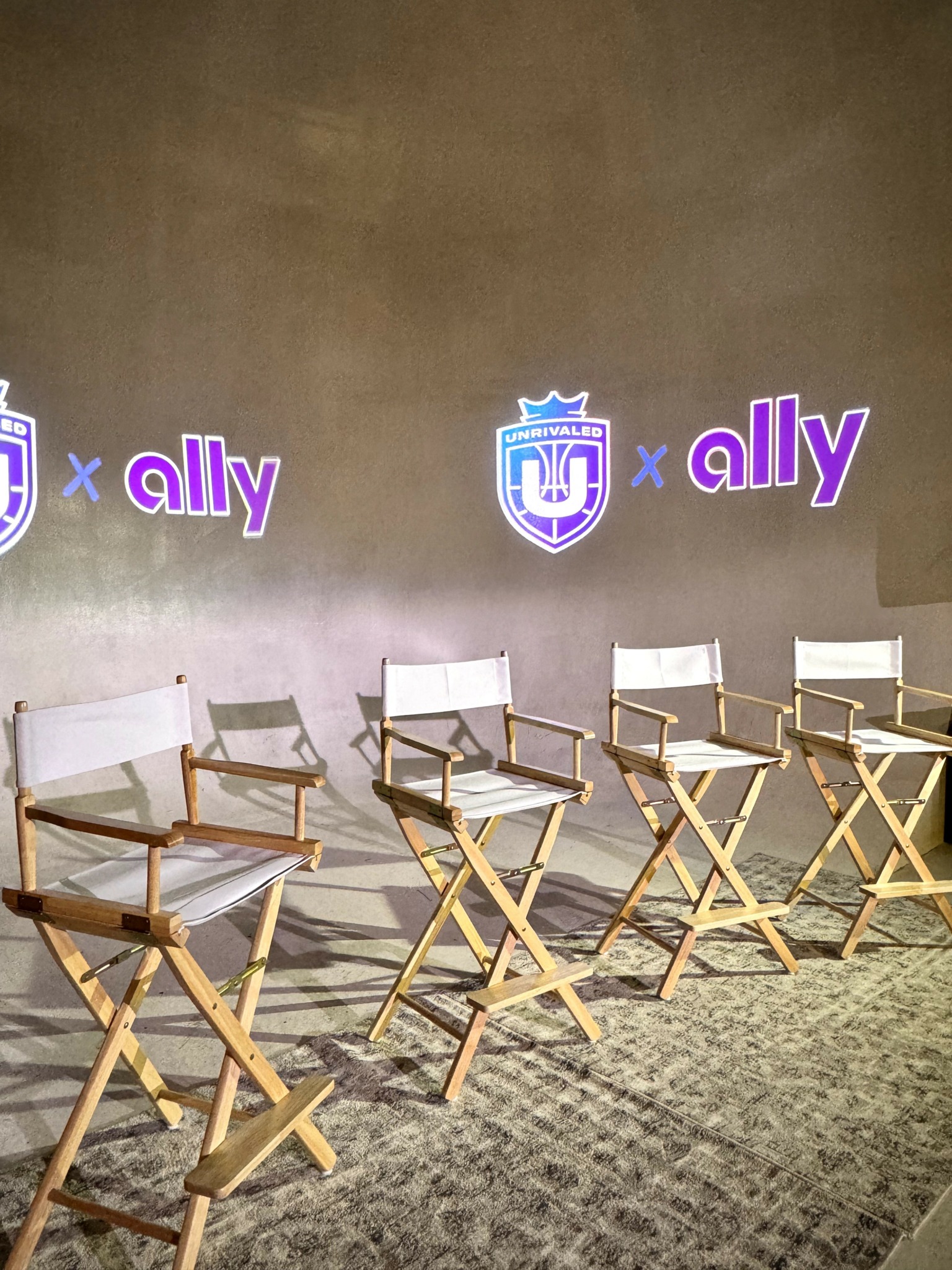 Six empty director's chairs with white seats and wooden frames are arranged in a row on a carpeted floor, with a wall displaying logos and the word 'ally' in the background.