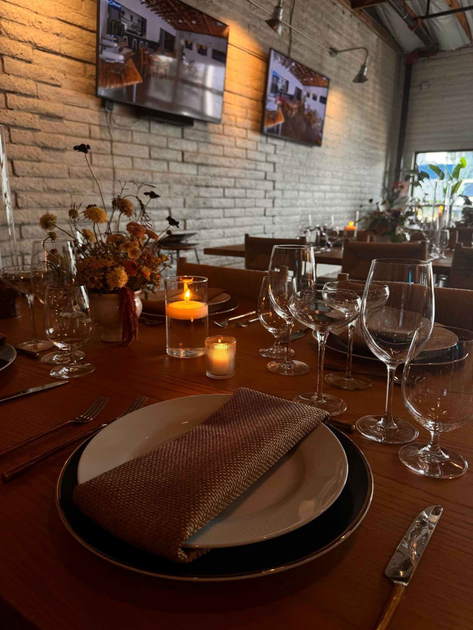 Dining table set with wine glasses, candles, and a plate with a napkin, in a cozy restaurant with brick walls.