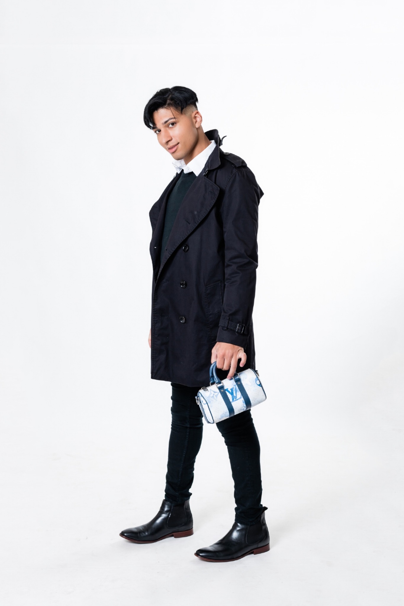 Young person standing, wearing a dark coat, holding a small bag, against a white background.