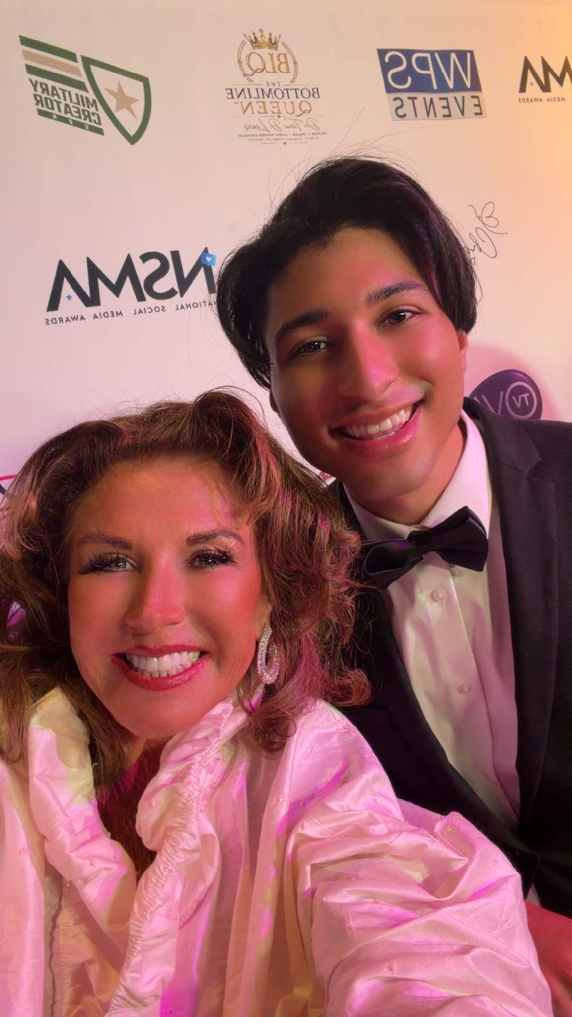 Two people smiling, one with curly hair wearing a pink outfit, the other with short dark hair in a tuxedo, in front of a backdrop with logos.