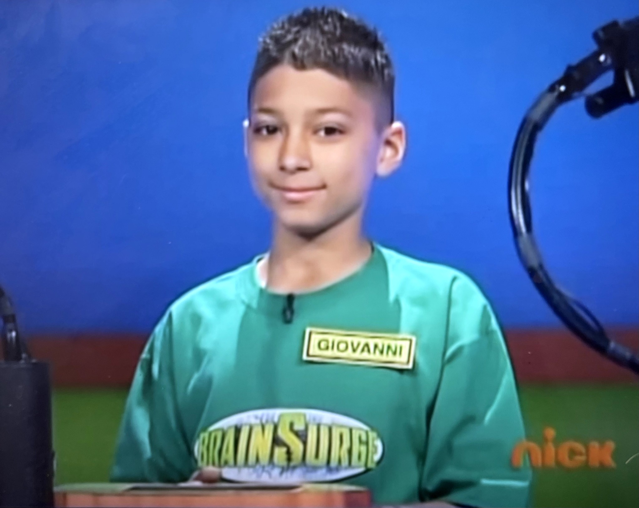 Young boy with short dark hair wearing a green shirt with a name tag and a logo, standing in front of a blue background.