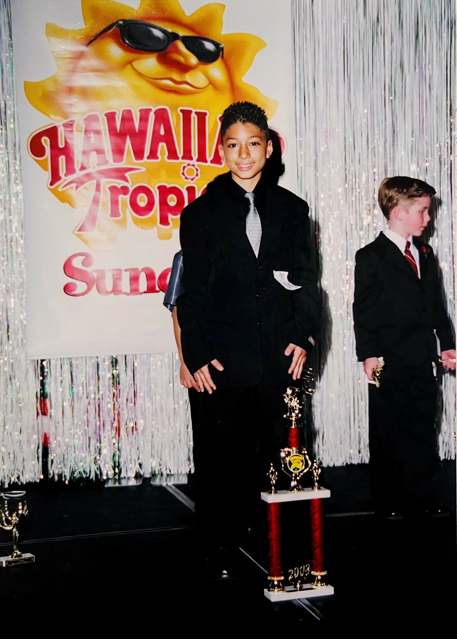 Two children in formal attire standing on stage with a Hawaii-themed backdrop and a trophy in front of the girl.