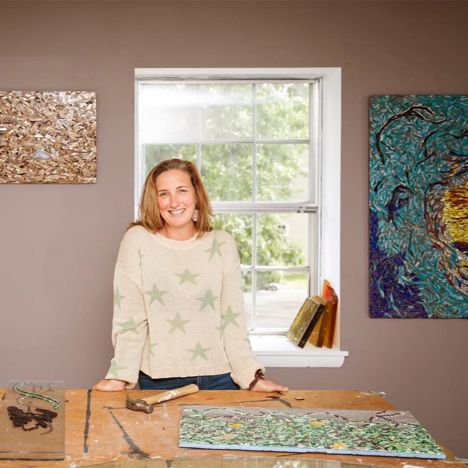 Smiling woman standing behind a wooden table with artwork, in front of a window with trees outside.