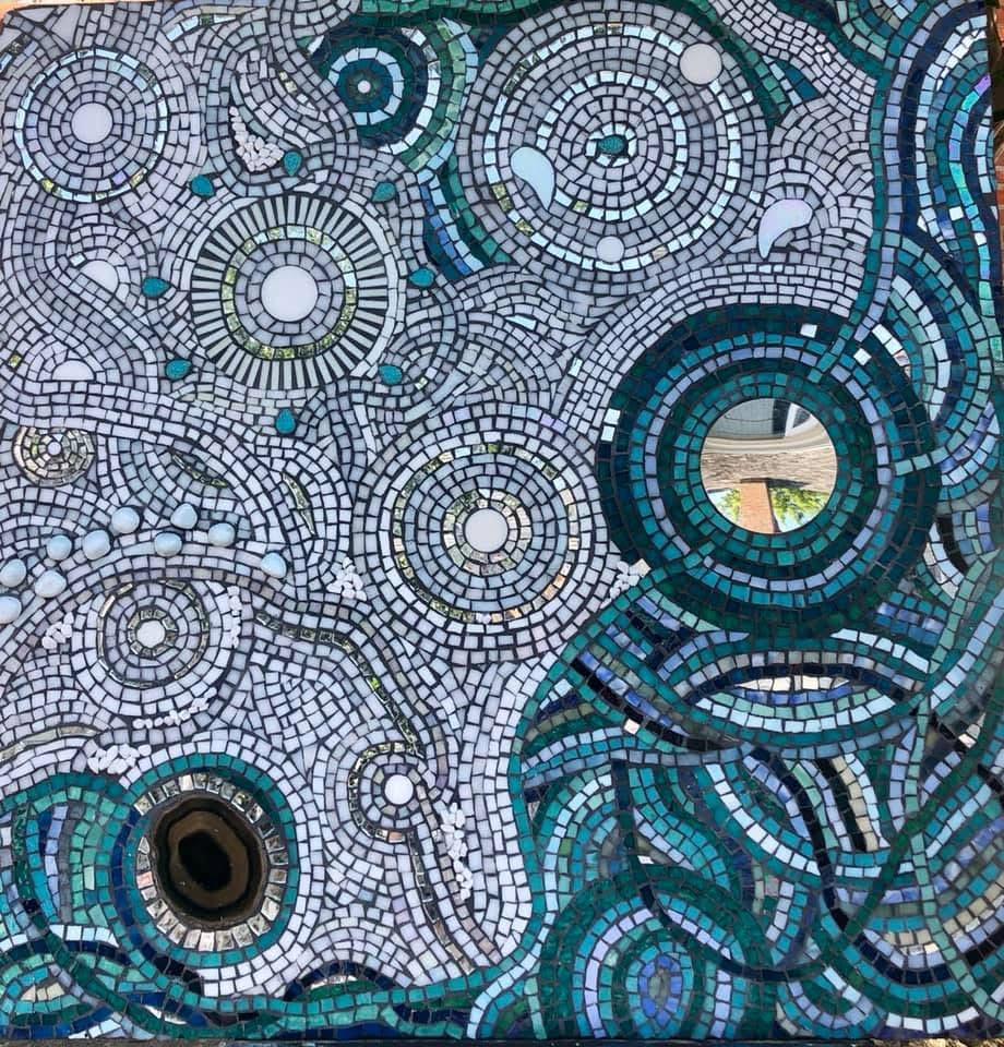 Colorful mosaic with circular patterns and a small opening showing a landscape, composed of small tiles in blue, green, and white.