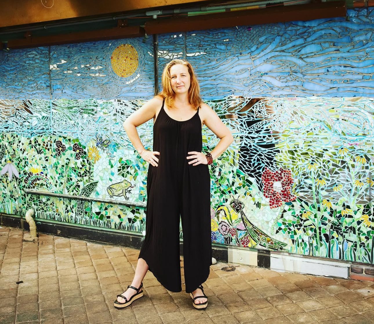 Woman standing with hands on hips in front of a colorful mosaic mural, smiling, wearing a black jumpsuit and sandals.