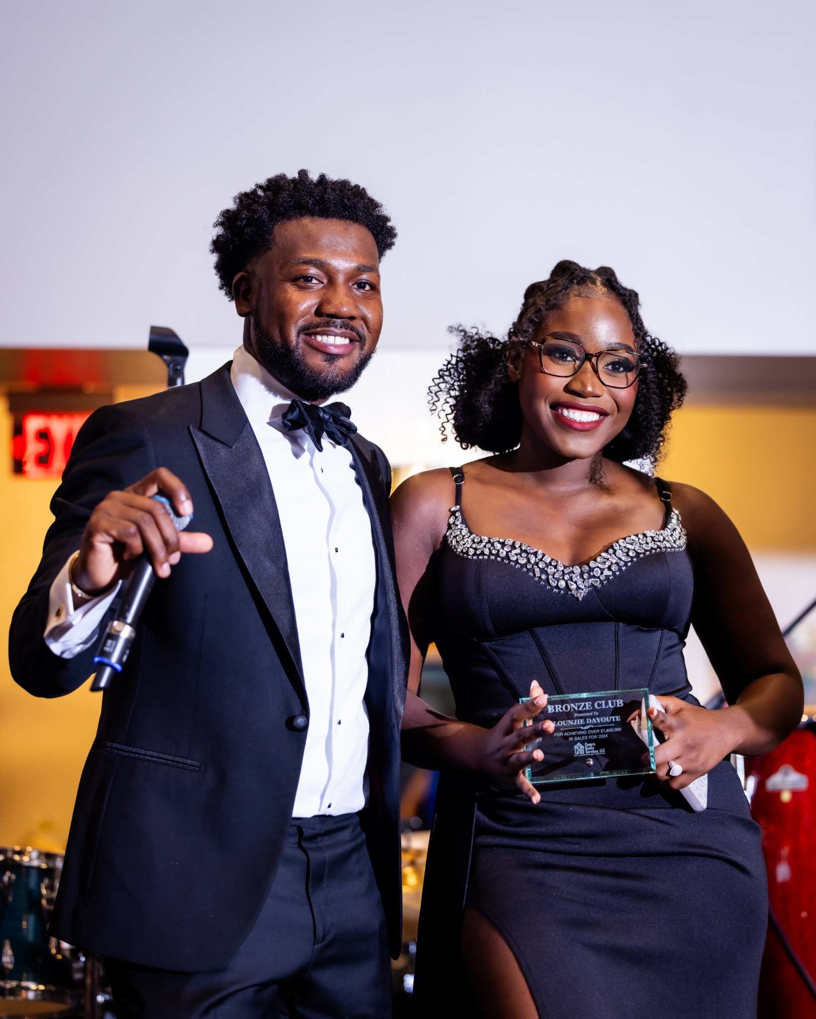 Man and woman in formal attire smiling, holding awards, standing indoors with a plain background.