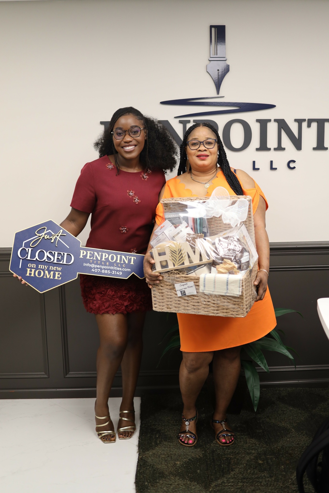 Two women stand indoors holding signs and a basket, smiling in front of a wall with a logo. One woman wears a red dress, the other an orange dress.