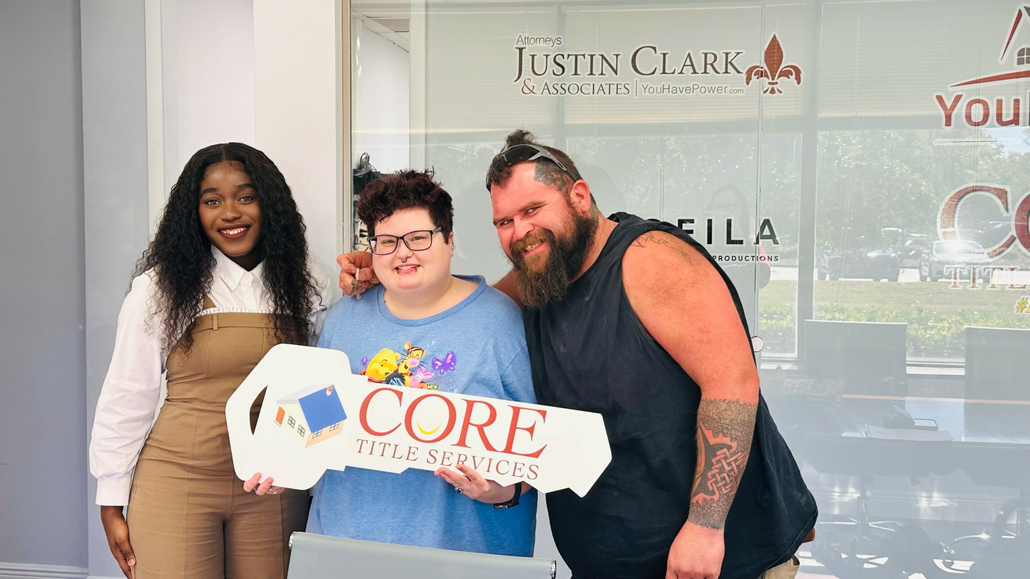 Three people smiling indoors, holding a large key-shaped sign that reads 'CORE TITLE SERVICES'.
