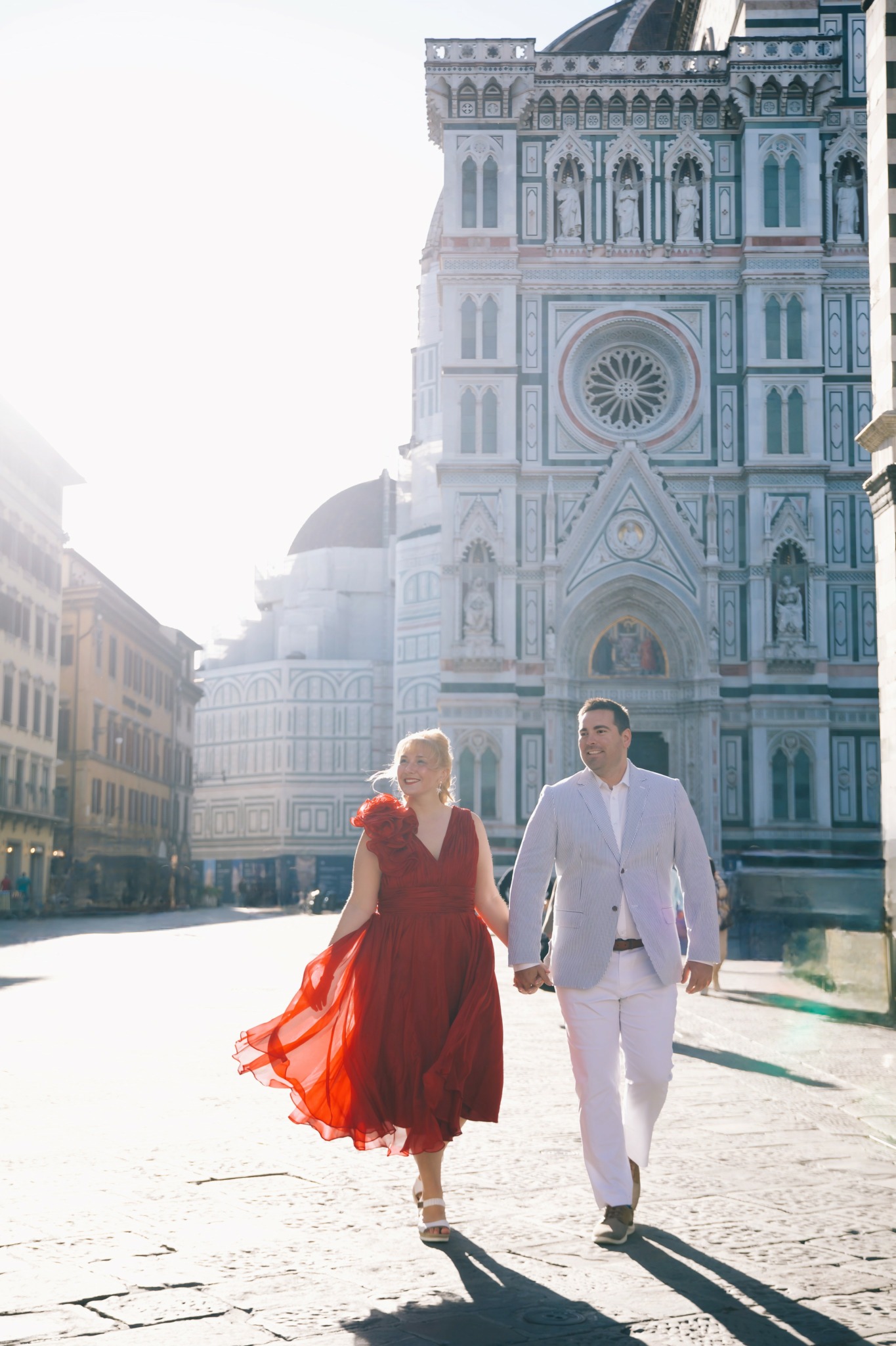 A couple walking hand in hand in front of a historic cathedral, sunlight shining behind them, woman in red dress, man in white suit.