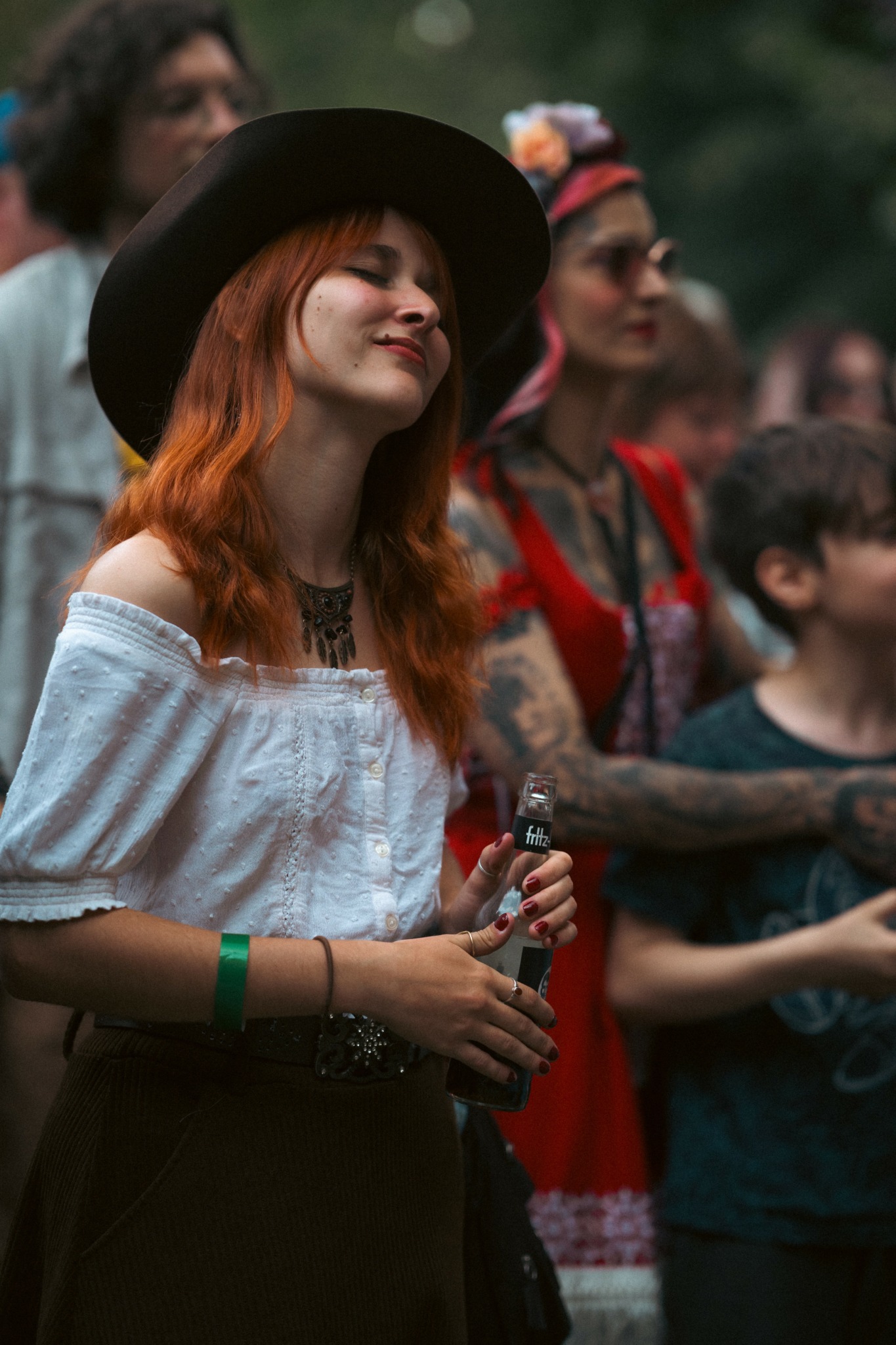Young woman with red hair, wearing a wide-brimmed hat and white off-shoulder top, smiling at an outdoor event.