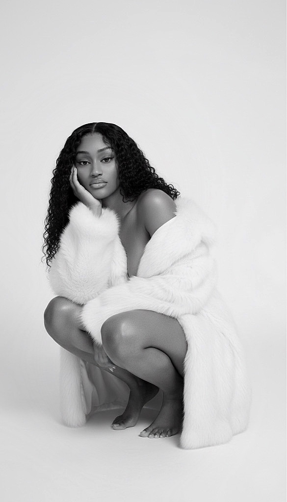 Woman with curly hair crouching, wearing a fluffy coat, against a plain background.