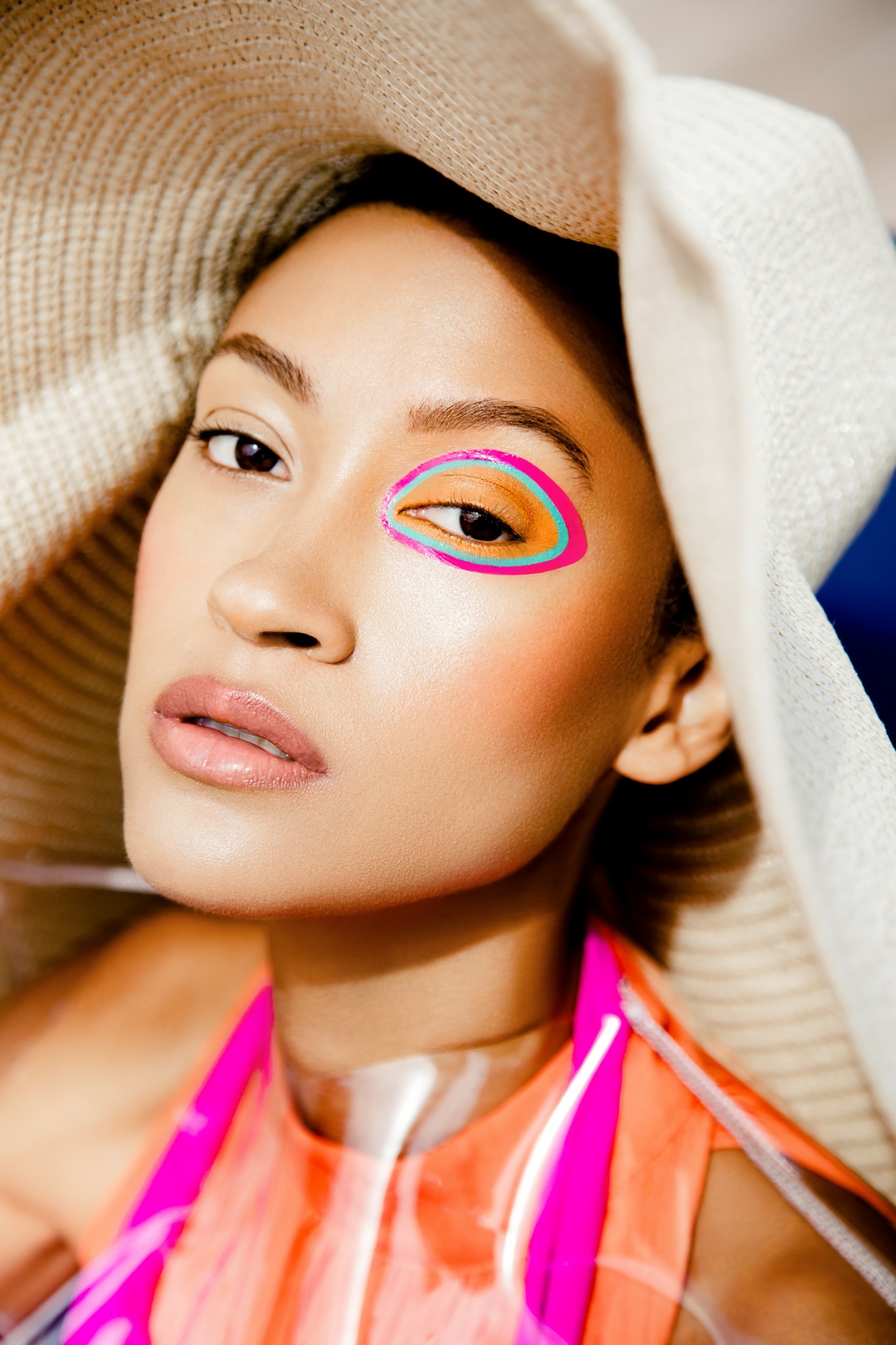 Close-up of a woman with colorful eye makeup and a large hat, looking at the camera.