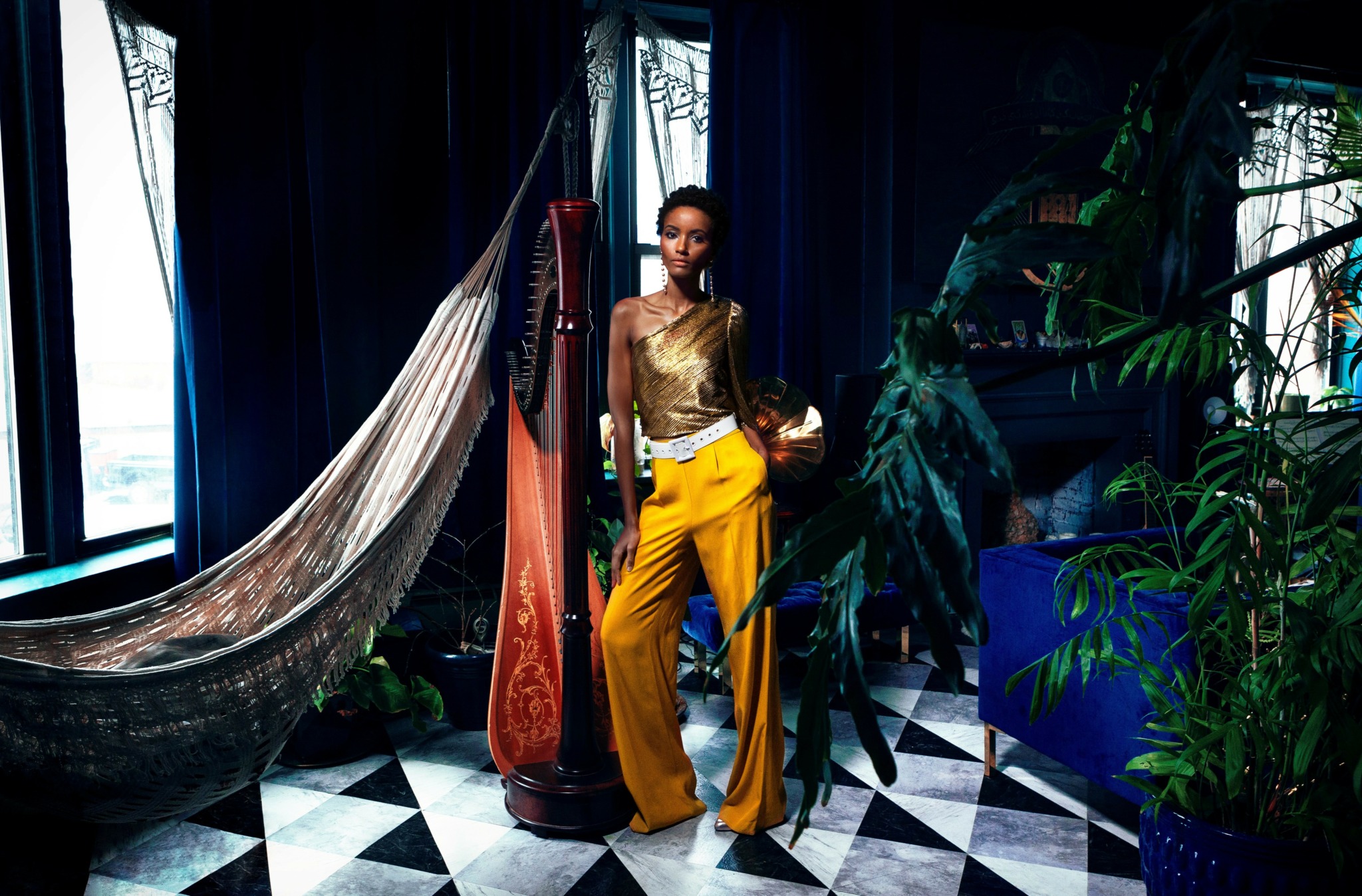 Woman in yellow pants and gold top standing near plants and a hammock in a room with black and white checkered floor.