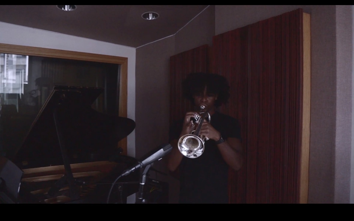 Person playing trumpet in recording studio with wooden panels and a piano nearby.