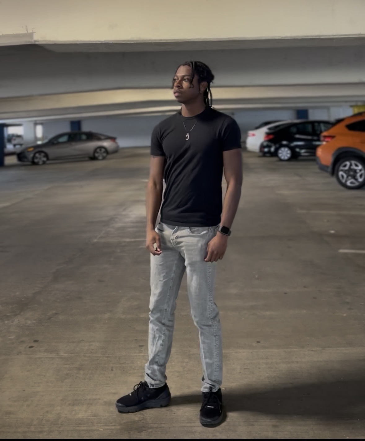 Young person standing in a parking garage, wearing a black t-shirt, light jeans, and black shoes, looking to the side.