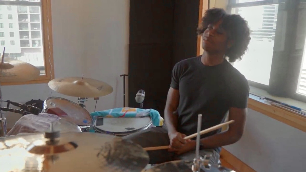 Person with curly hair holding drumsticks in a room with windows and musical instruments.