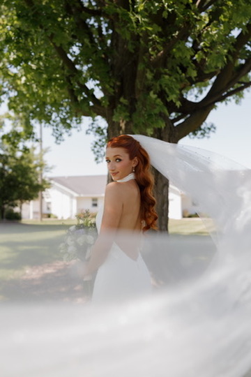 Woman with long hair standing outdoors under a large tree, with a white veil flowing around her, bright sky background.